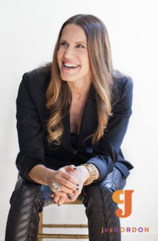 Woman sitting on low stool in front of a white wall. She is wearing a long sleeve black shirt leaning forward and laughing. She has long brown hair and several silver skull rings on her fingers. The right is over the left hand.