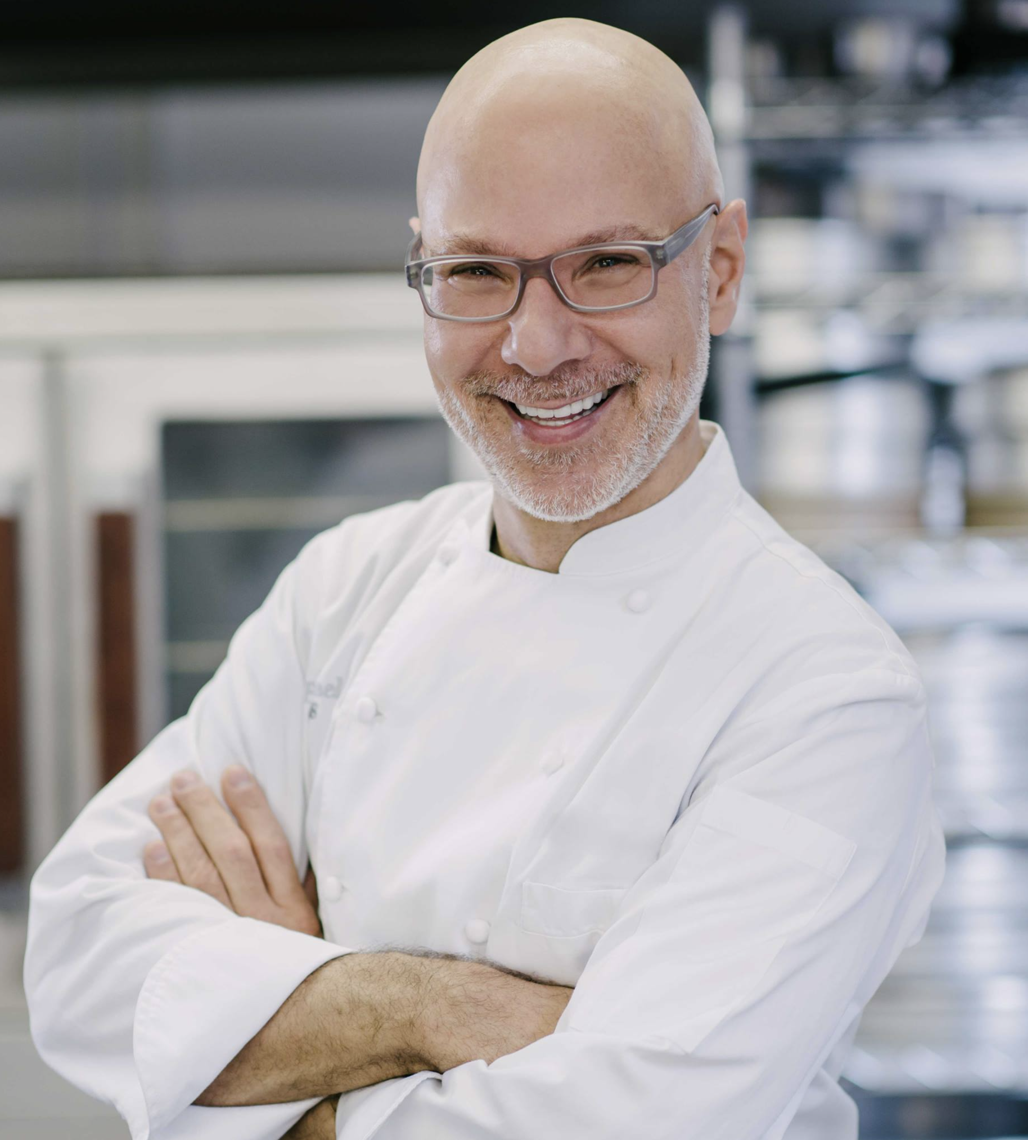 Close-up photo of a balding male baker in white apron wearing glasses outside crossing his arms and smiling at the camera.