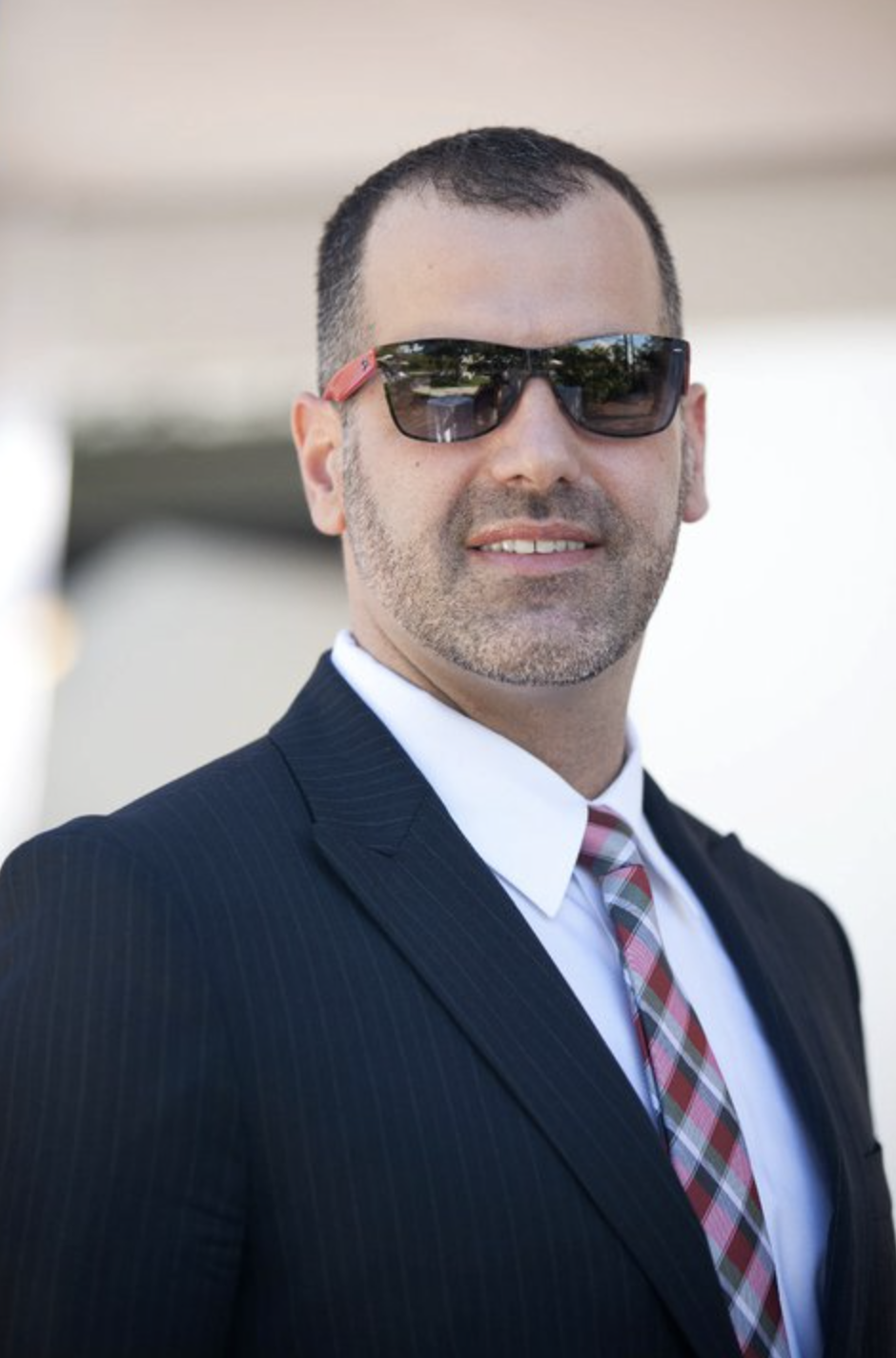 Man in sunglasses looking at camera wearing a blue jacket with white button down shirt, striped white and maroon tie.