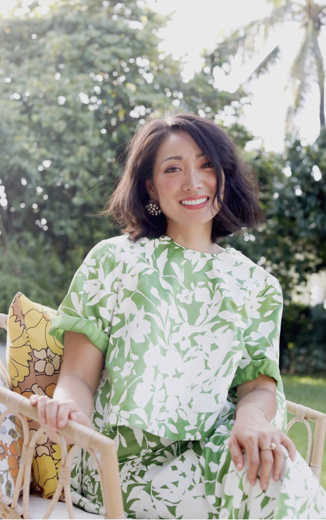 Asian woman with short black hair sitting on a lounge chair outside in a backyard. She is wearing a green dress with white flowers