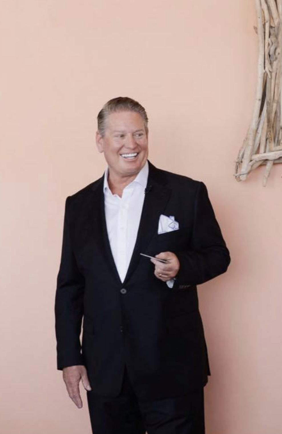 Three quarter photo of a man standing against a moave colored wall. The man is wearing a black dinner jacket with a button down white shirt. He has a white pocket square and is looking off to his left and smiling.