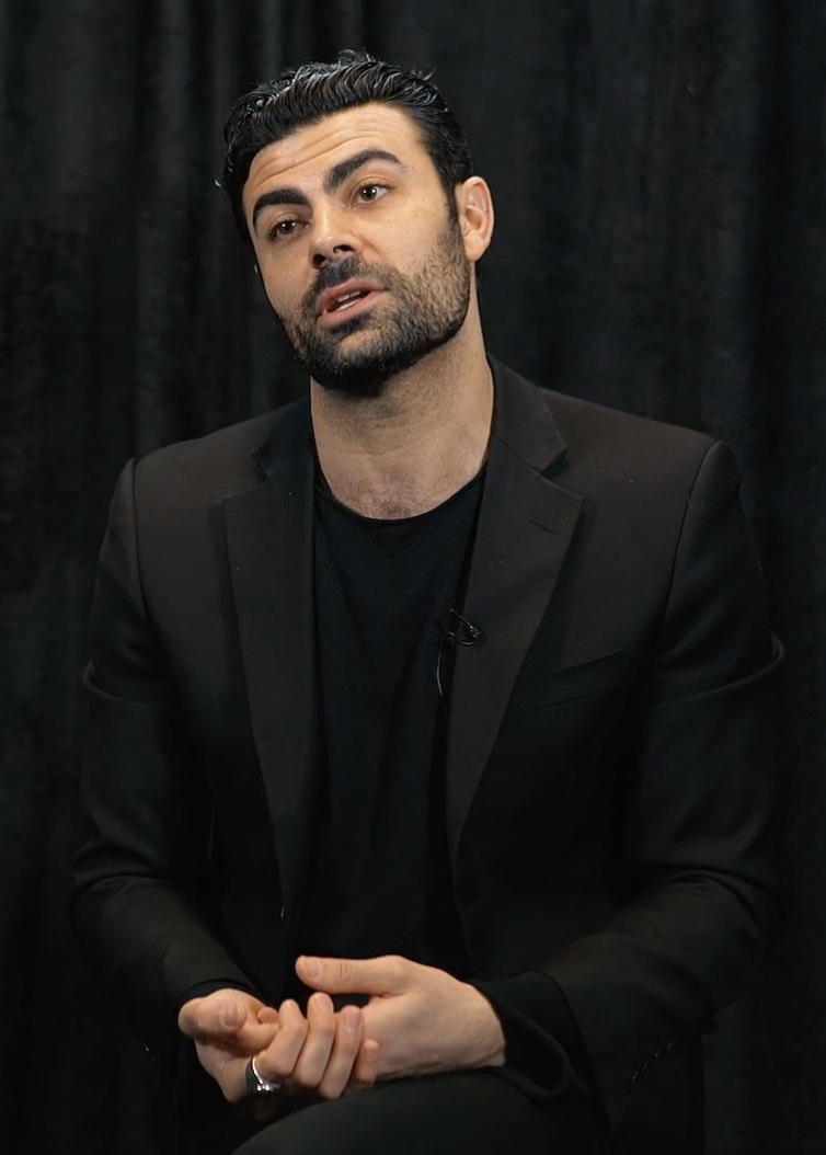 Man with full beard and a mustache being interviewed wearing a black jacket and black crew neck shirt in front of a black background.