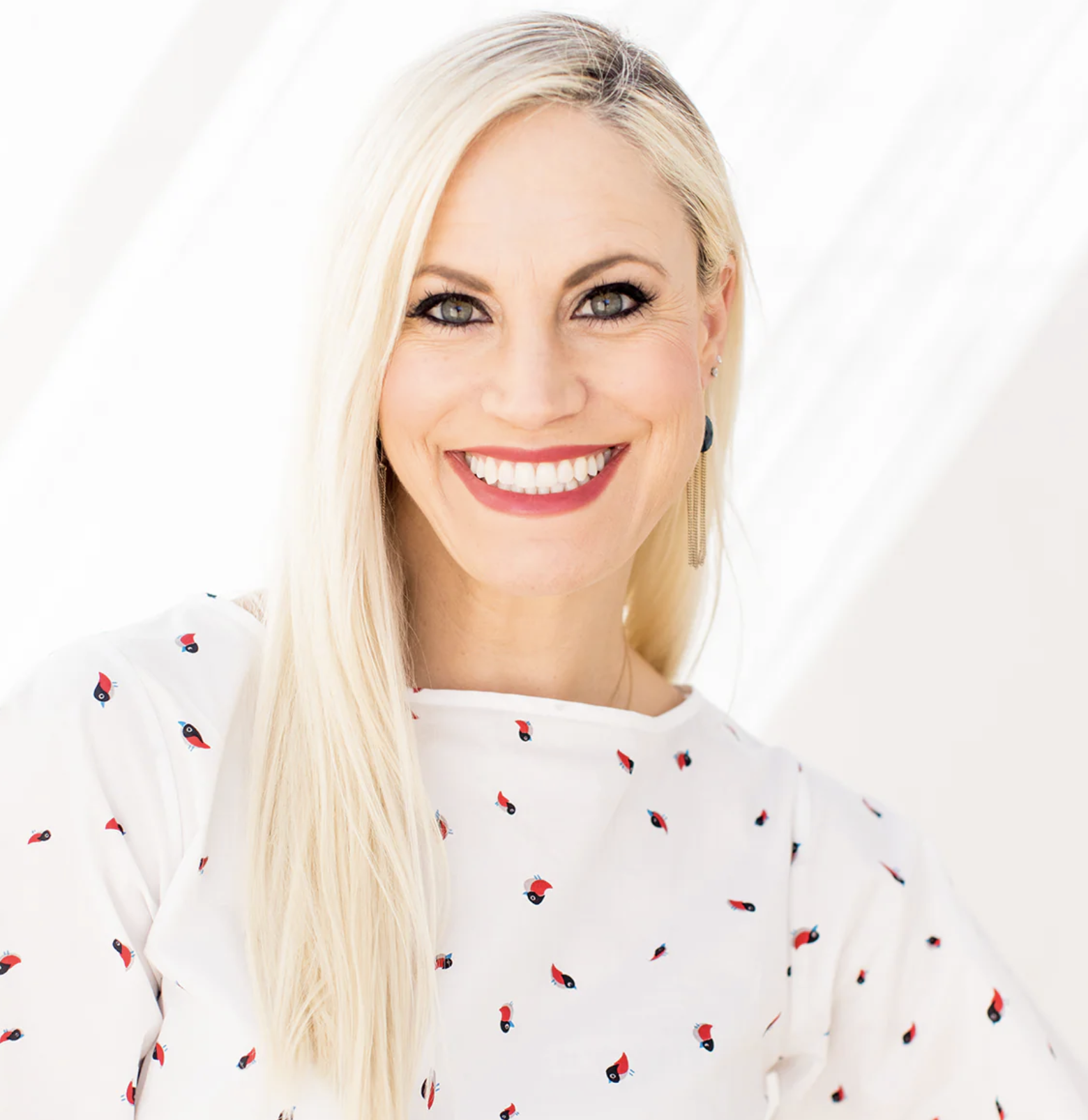 Long blonde haired caucasian woman that's also black roots. She's smiling at camera and it's a close-up photo. She's in front of a white background wearing a white shirt with red dots.