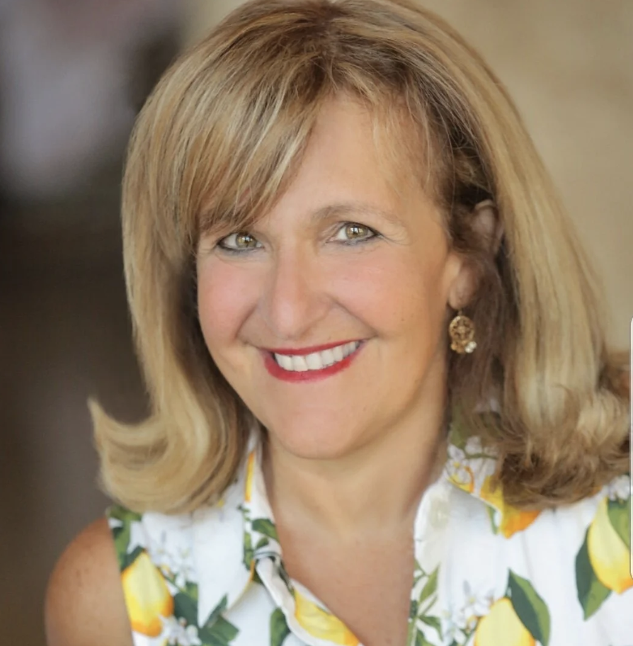 dirty blonde haired woman smiling at the camera. It's a close-up photo and she's wearing makeup and a white, dress with green and yellow flowers.